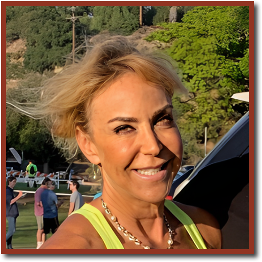 A smiling woman with short blonde hair stands outdoors in a sunny setting. She’s wearing a light green tank top and a necklace, with her hair blowing slightly in the wind. In the background, there are a few people gathered in what appears to be a park or an event, with trees and hills visible in the distance.